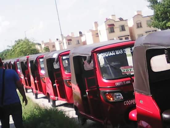 A row of red auto rickshaws parked on the side of a street with multi-story residential buildings in the background under a clear sky.