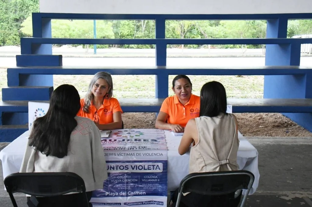 Two women in orange shirts sitting at a desk with informational banners, talking to two other women whose backs are to the camera