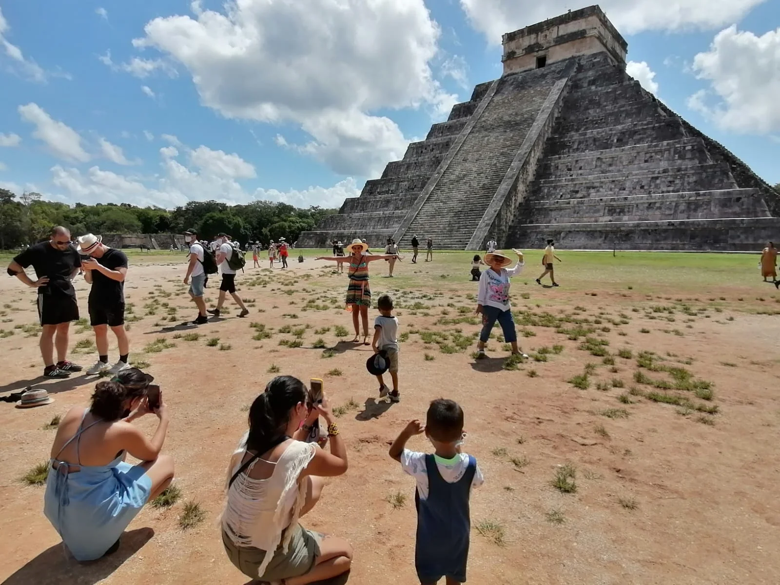 Tourists enjoying and taking photos at the base of the El Castillo pyramid at Chichen Itza under a sunny sky with scattered clouds.