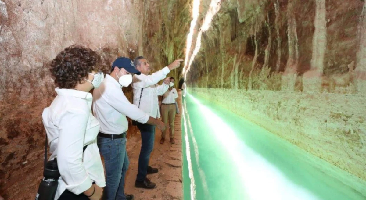Visitors observing a subterranean water channel illuminated by green light inside a cave-like structure while wearing masks and pointing out features.