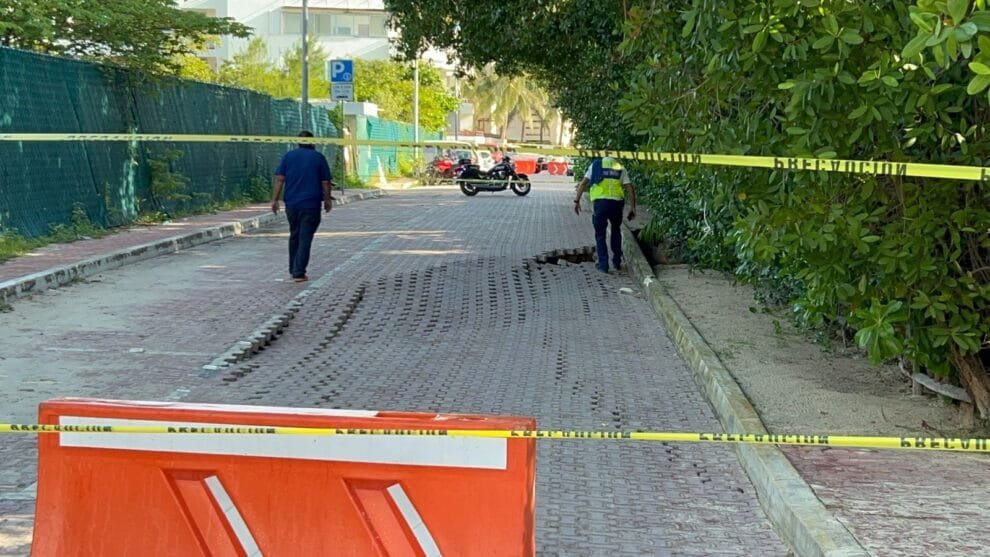 Two uniformed officers inspect a cordoned-off area on a shaded street, with a red barrier and yellow crime scene tape in the foreground.