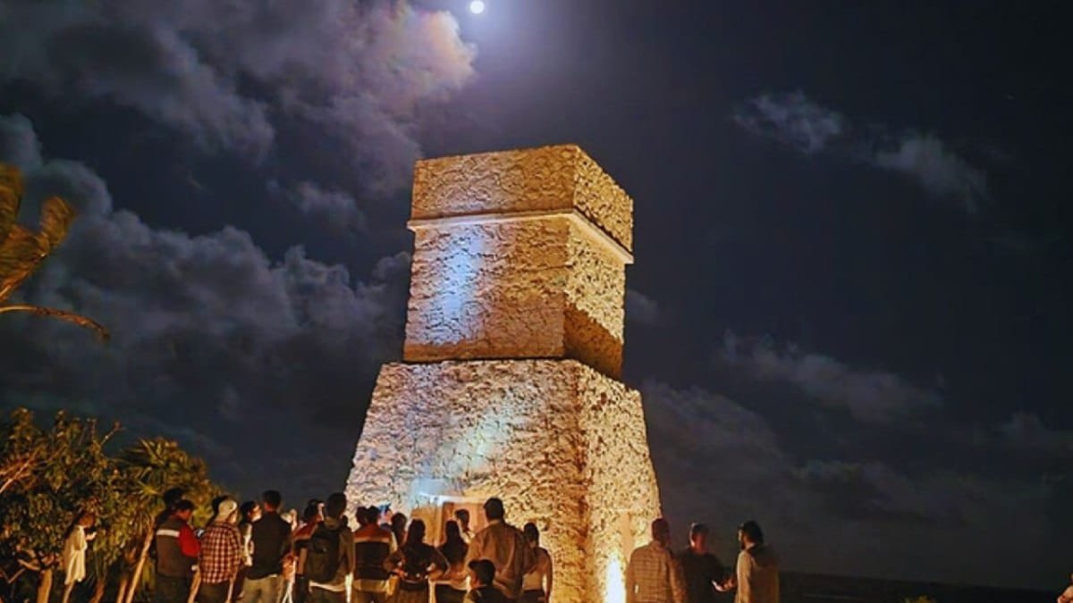 People gathered at night around an illuminated ancient stone tower with the moon partially visible in the sky.