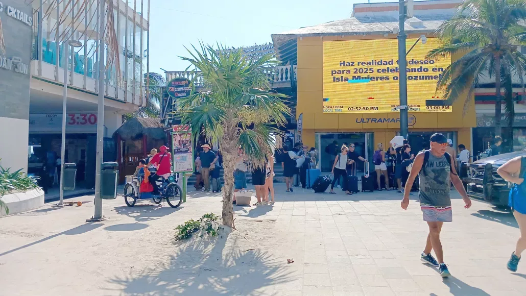 A sunny tropical street scene with palm trees, tourists, and various shops, including an Ultramar ferry office.