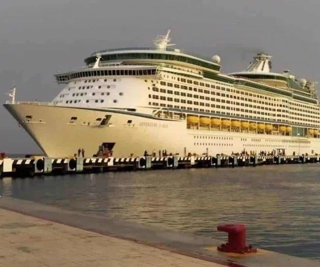 A large white cruise ship, Adventure of the Seas, docked at a sunny port with a clear blue sky in the background and a red bollard in the foreground.