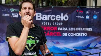 A man wearing an Animal Equality t-shirt speaking into a microphone at an outdoor event with banners advocating against dolphin cruelty in the background