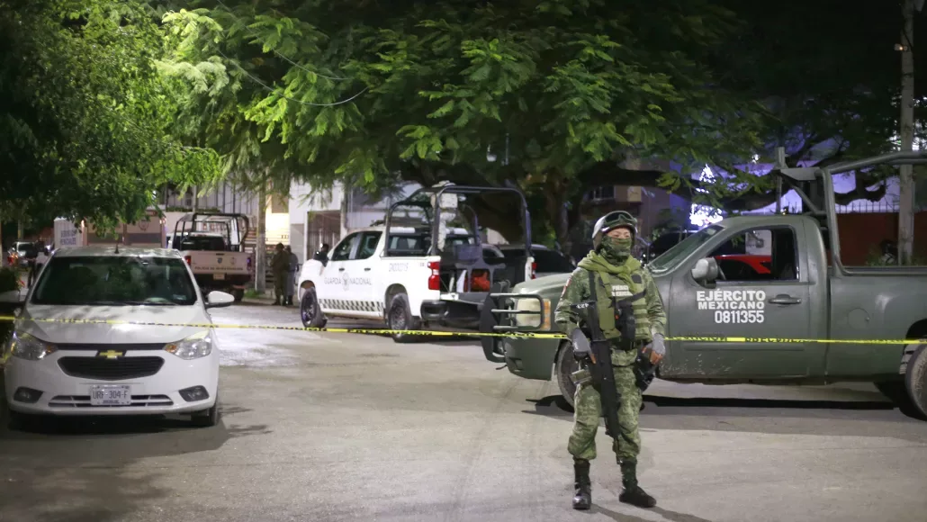 A military personnel in camouflage stands guard at a nighttime security operation, with marked vehicles and caution tape around an urban street scene.$# CAPTION