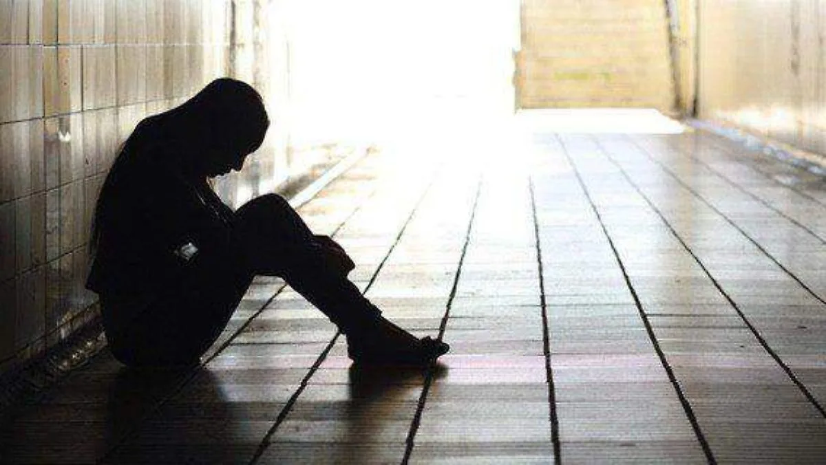 A silhouette of a person sitting head down in a dimly lit corridor, illuminated by a bright light at the end, conveying a sense of loneliness and contemplation.