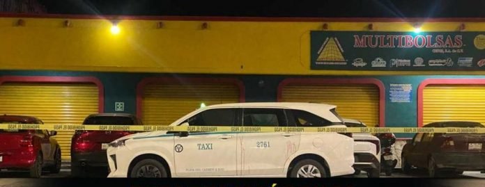 A white taxi alongside other vehicles behind police tape at a nighttime crime scene in front of a yellow building labeled "Multibolsas".