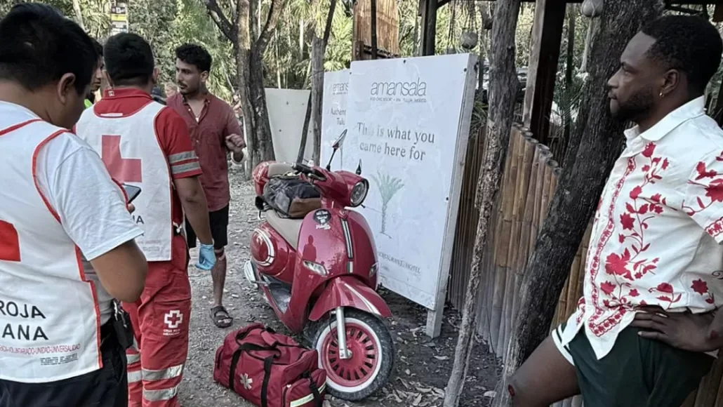 Three men in Red Cross uniforms discussing near a red scooter, with another man in casual clothing observing, in a rustic outdoor setting.