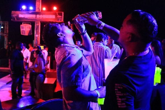 A person drinking from a bottle at an outdoor nighttime event with other people and colorful lights in the background